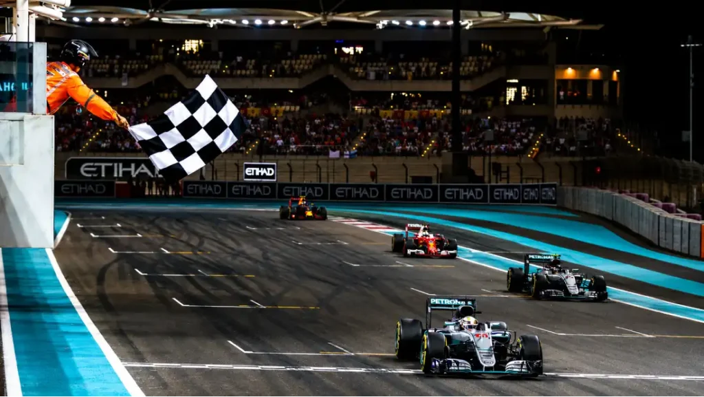 A race official waving a checkered flag at the finish line as a Mercedes F1 car crosses the grid at night during a Grand Prix.